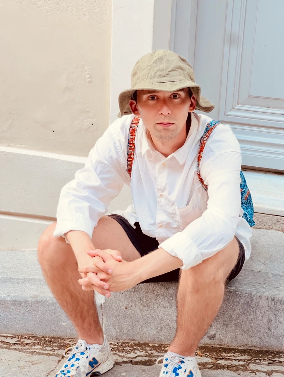 Elias Grünewald Dykert in a bucket hat and white shirt, sitting on a stone step by a pale blue door